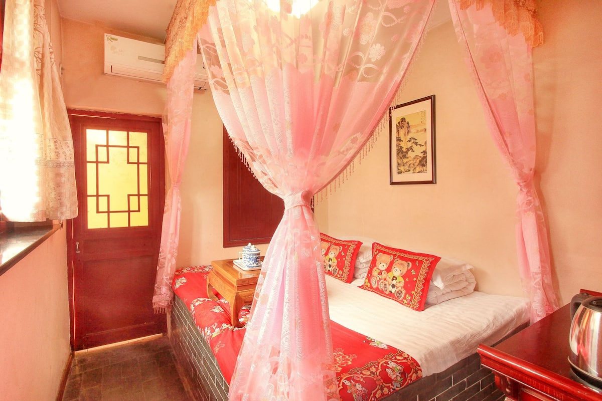 A traditional room with a bed, a wooden nightstand, a red and white patterned bedspread, and a pink canopy.