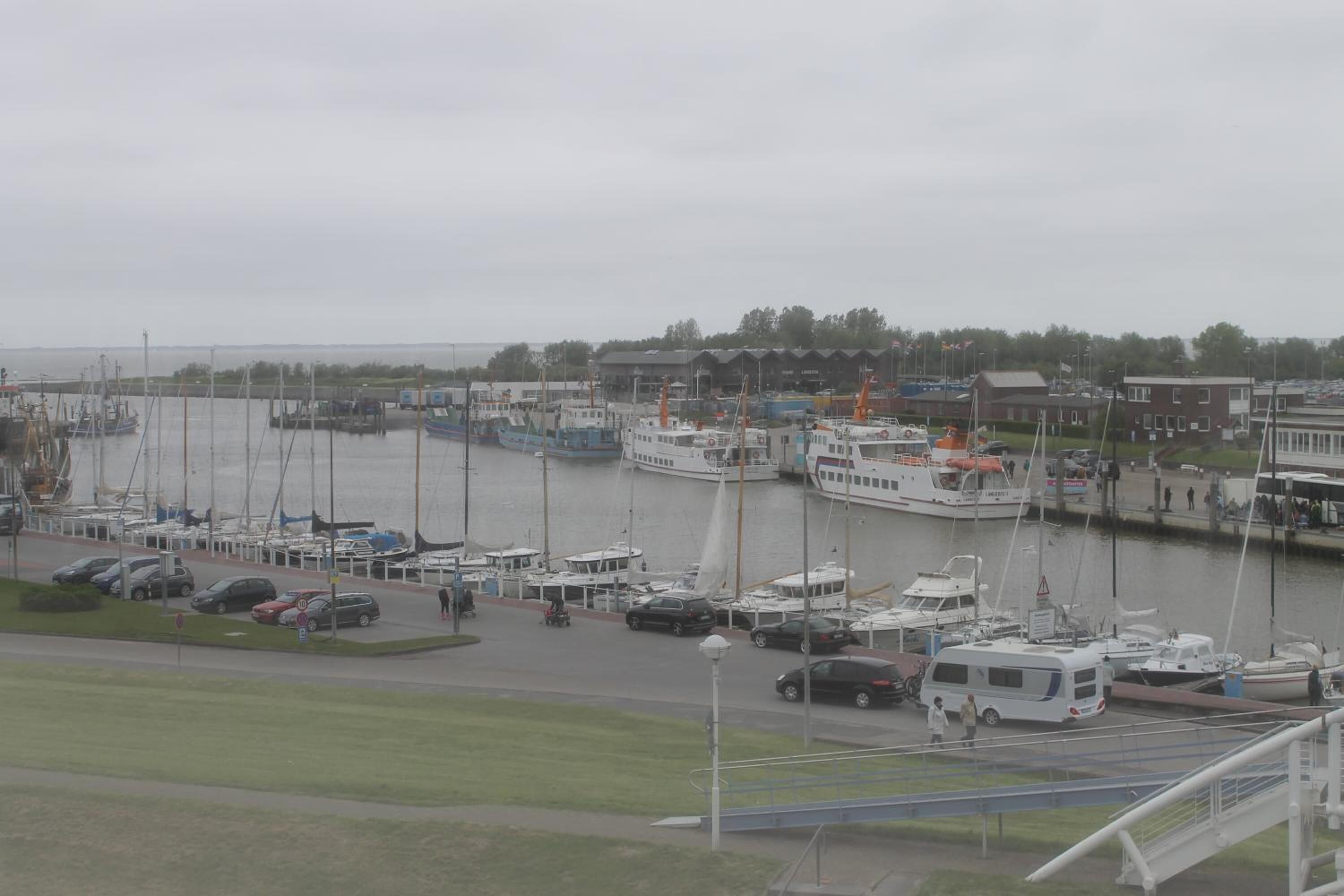 Un puerto deportivo con varias embarcaciones amarradas, un área de estacionamiento con automóviles y un edificio al fondo.