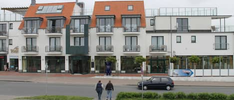 A row of modern buildings with balconies, a street in front, and a car parked on the side.
