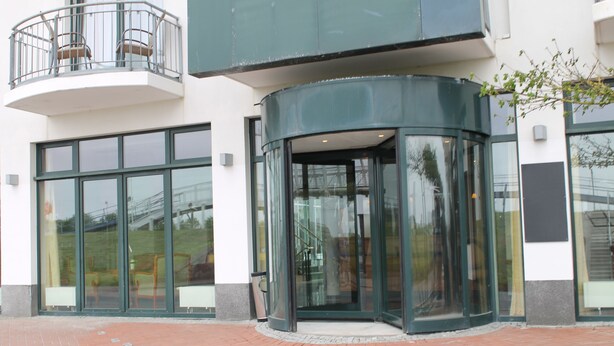 Un bâtiment doté de grandes portes vitrées, d’une entrée incurvée et d’un petit balcon orné de rampes en métal.