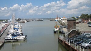 Un port de plaisance avec des bateaux amarrés le long d’une jetée incurvée, un petit bâtiment en briques et une rangée de voitures garées sur la route adjacente.