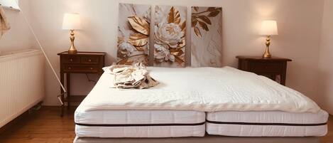 A bedroom with a bed, two bedside tables with lamps, a radiator, and a wall with floral artwork.