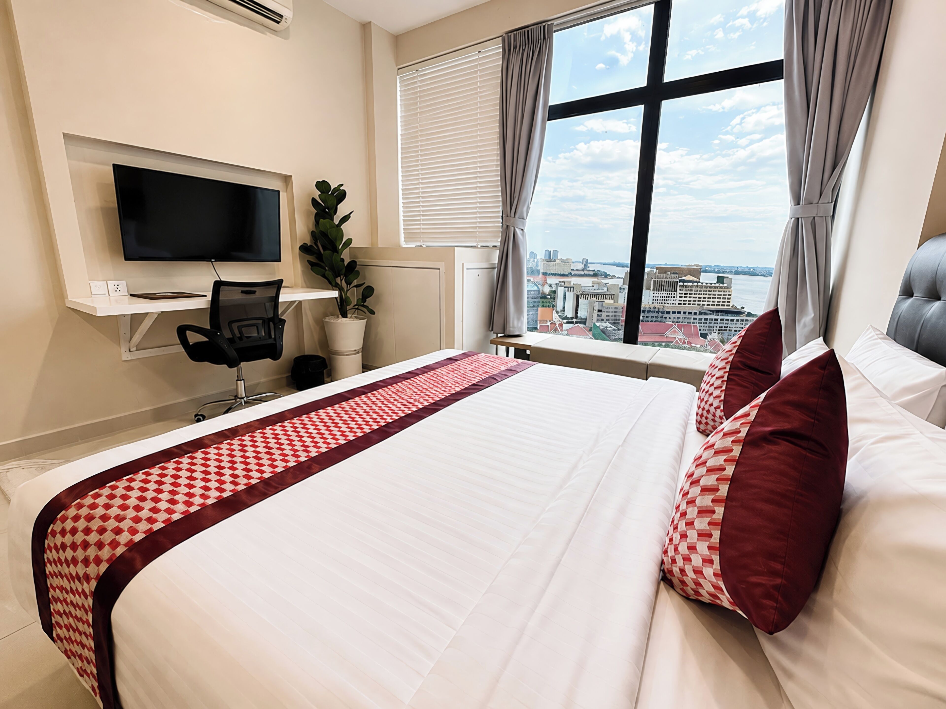 A hotel room with a bed, a desk with a chair, a television, a potted plant, and a window with a city view.