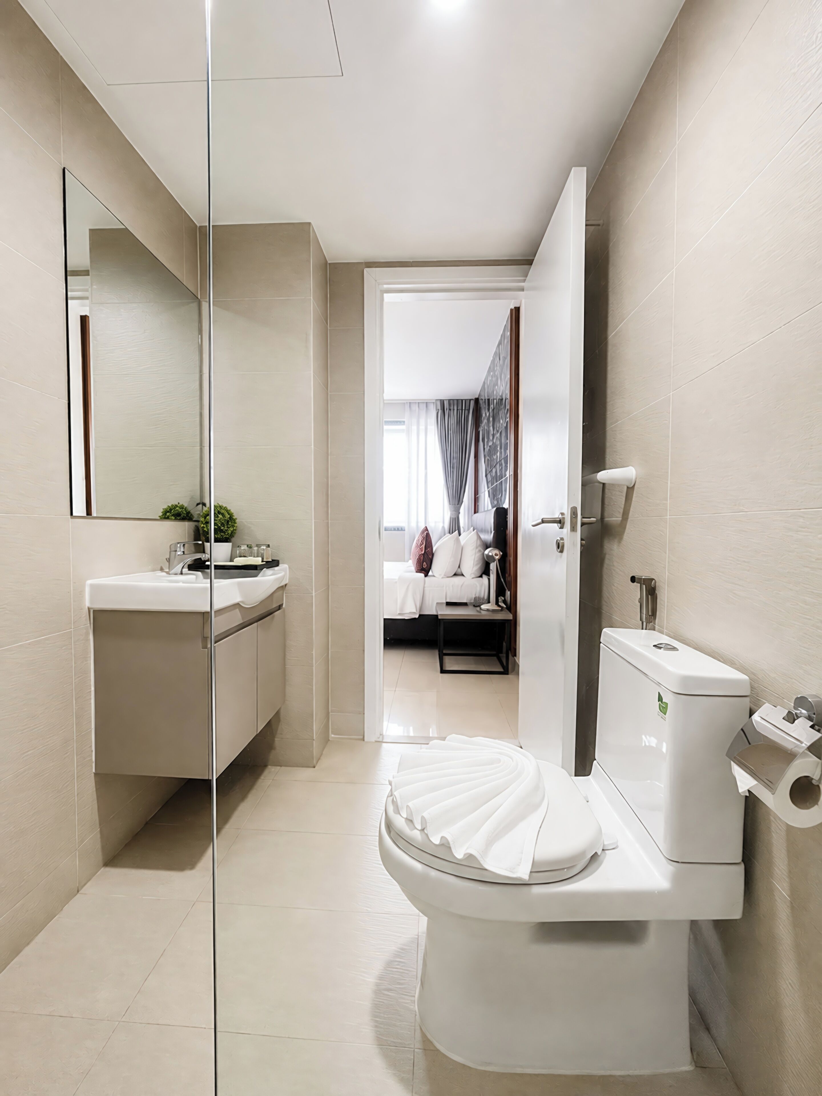 Un baño moderno con inodoro, lavamanos y espejo. Se ve una cama a través de una puerta abierta al fondo.