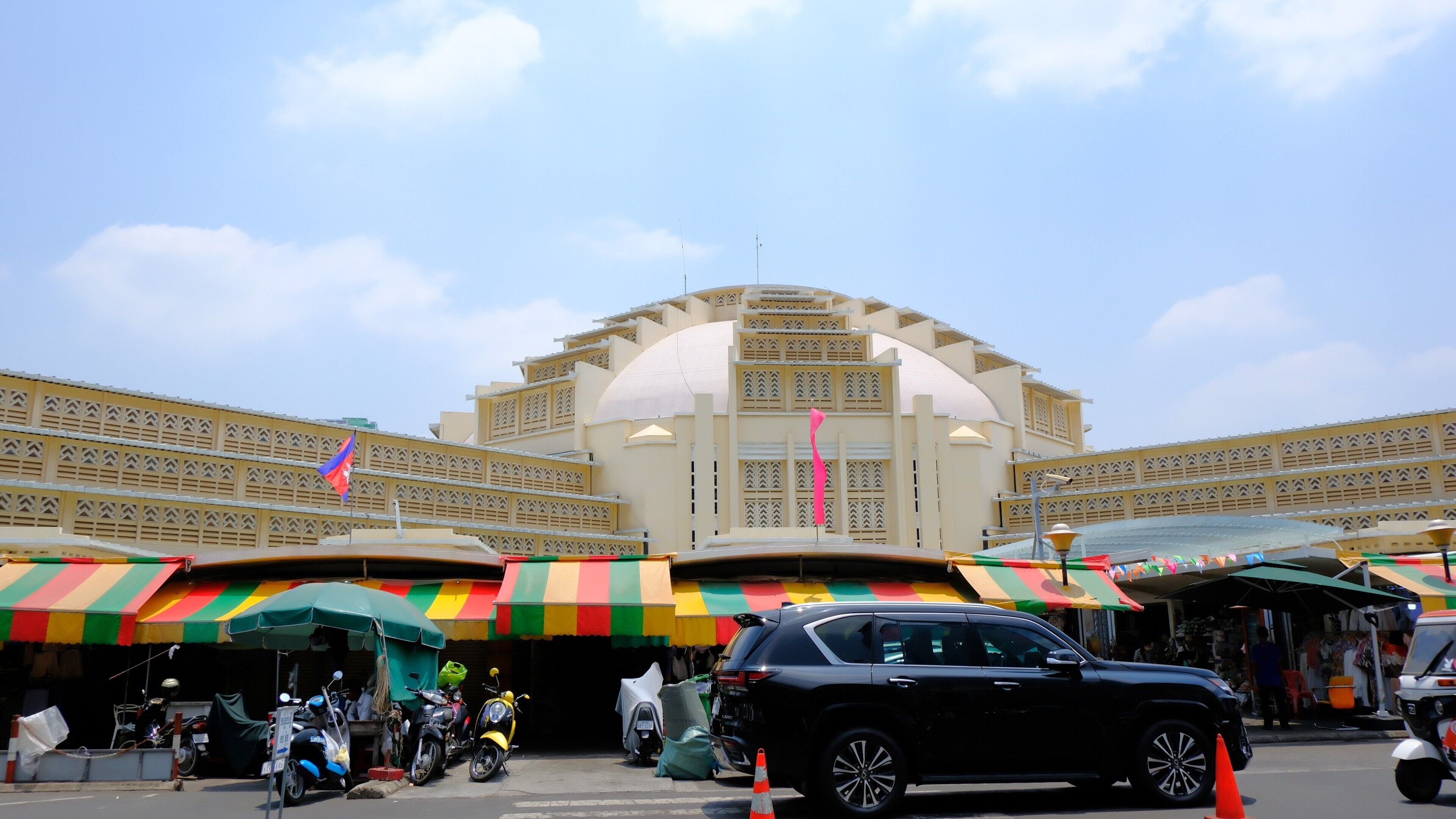 A large building with a dome and a flat roof, surrounded by colorful umbrellas and parked vehicles.