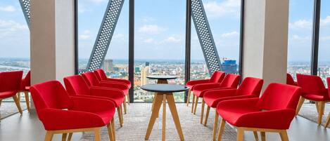 A conference room with red chairs arranged around a central table, large windows offering a cityscape view.