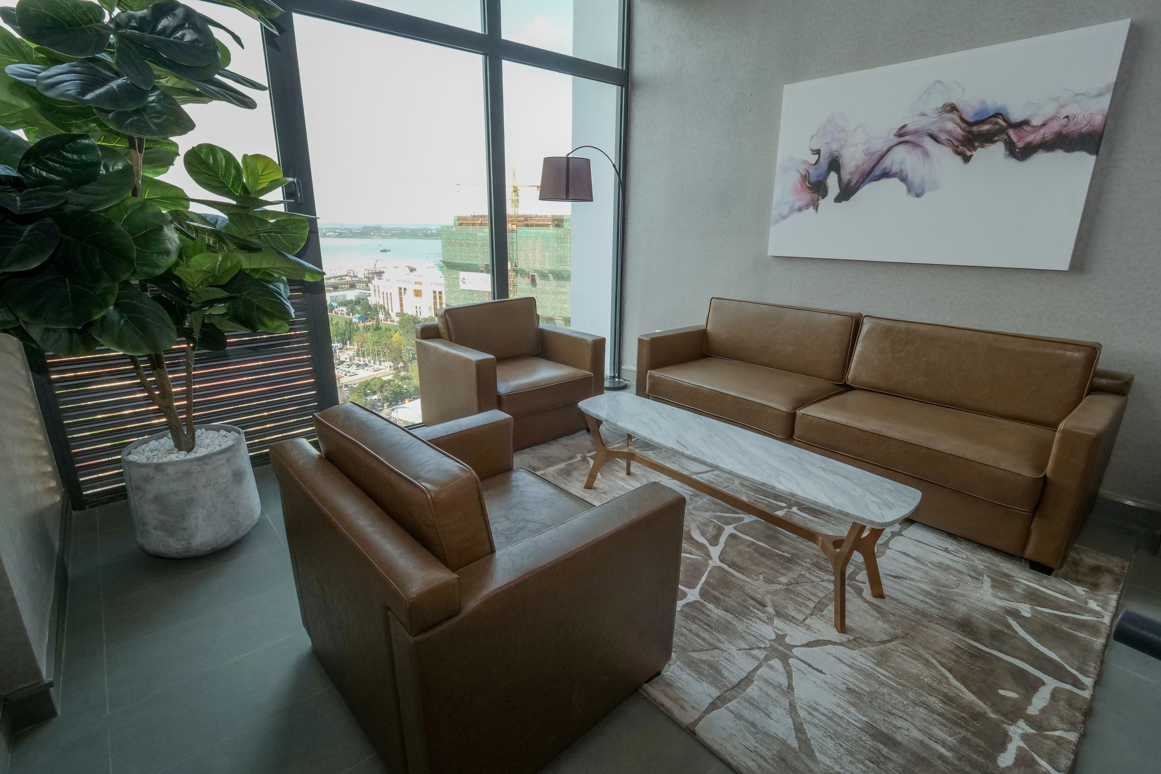 A modern living room with a brown leather sofa, a coffee table, and a large window offering a view of the cityscape.