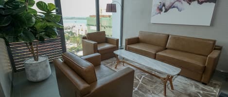 A modern living room with a brown leather sofa, a coffee table, and a large window offering a view of the cityscape.