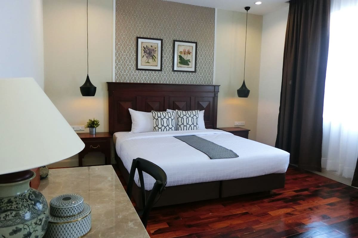 A neatly arranged hotel room with a large bed, a bedside table with a lamp, a small potted plant, and two framed pictures on the wall.