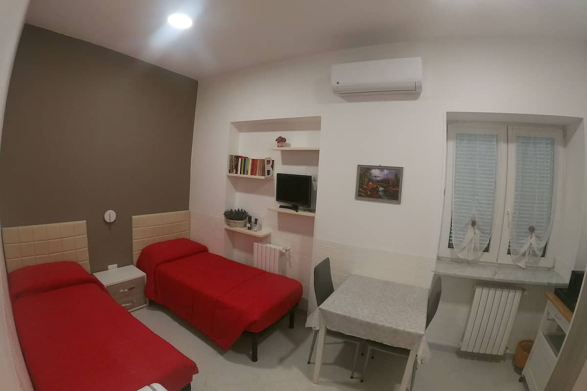 A small, well-lit room with two single beds, a desk with a chair, a radiator, and a window with blinds.
