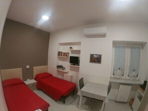 A small, well-lit room with two single beds, a desk with a chair, a radiator, and a window with blinds. - Ostello Villafranca (Pignola)