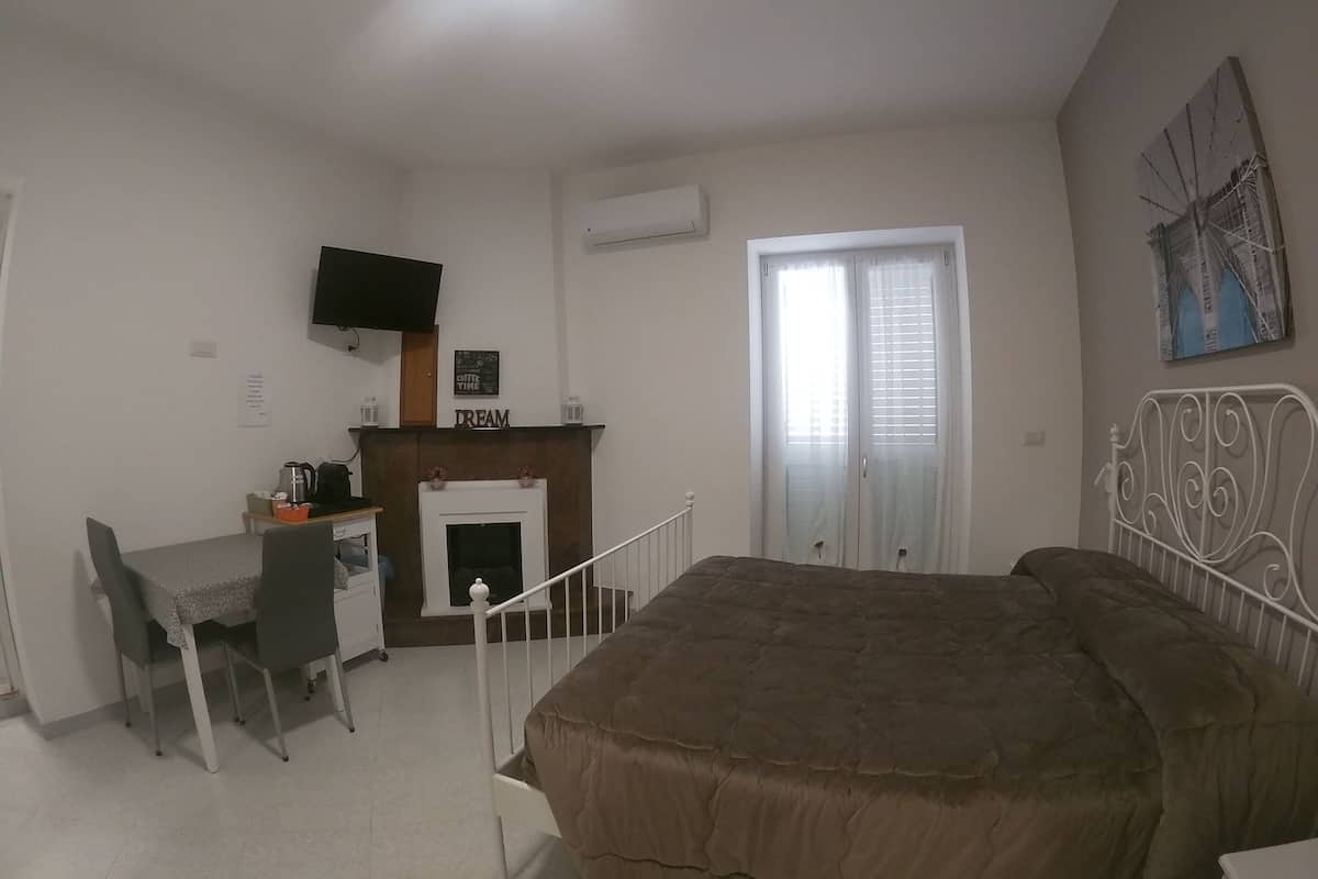 A bedroom with a bed, a desk with a chair, a fireplace, a television mounted on the wall, and a window with blinds.