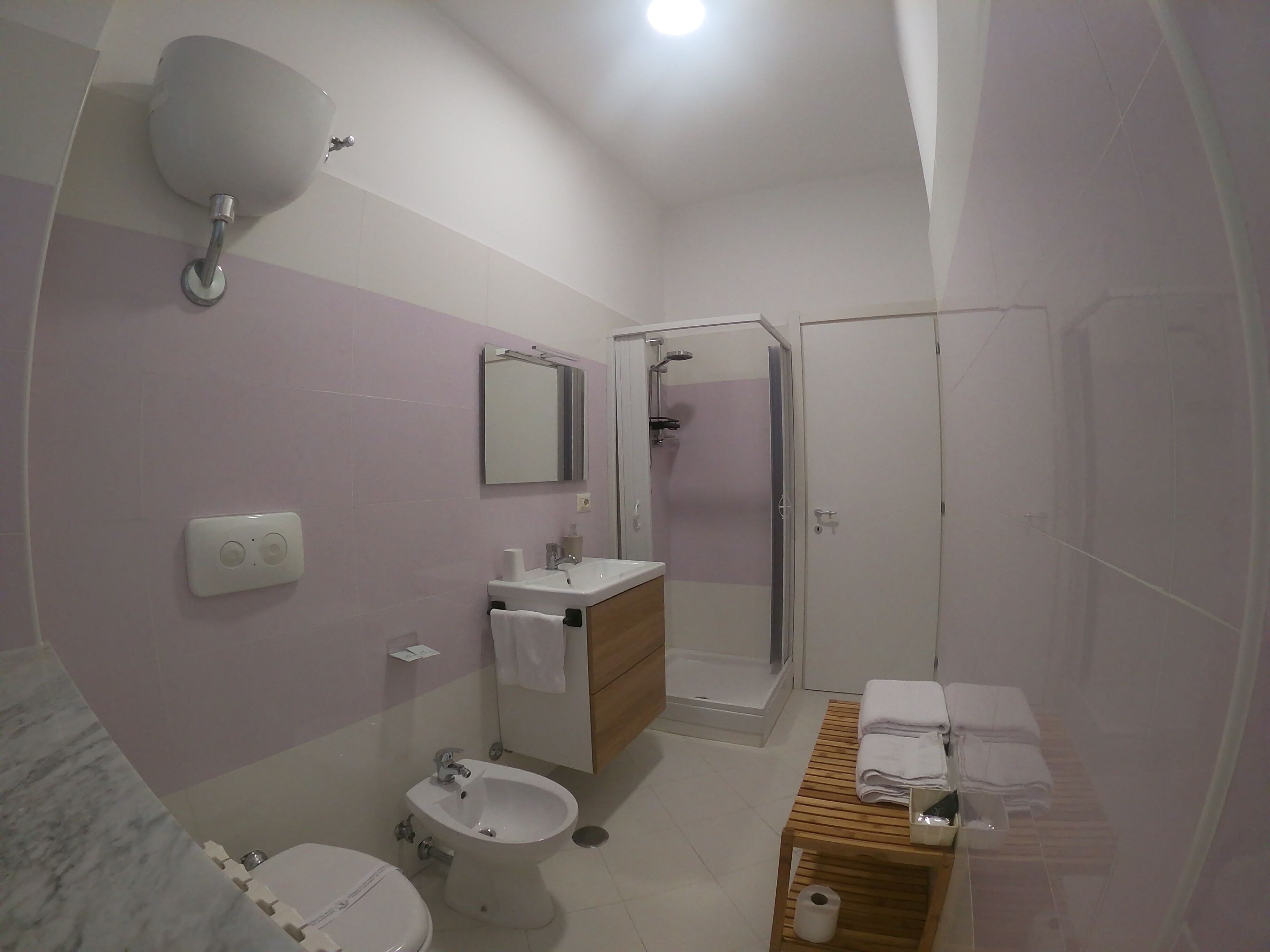 Bagno con toilette, bidet, lavandino, specchio, doccia e una panca di legno con asciugamani.