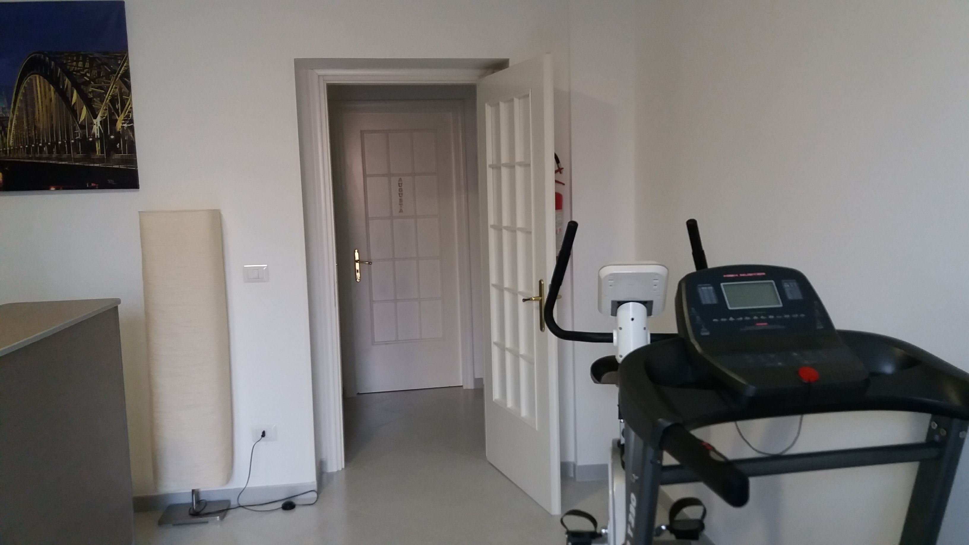Ein Zimmer mit Laufband, einer Tür zu einem anderen Raum und einem an der Wand montierten Lichtschalter.