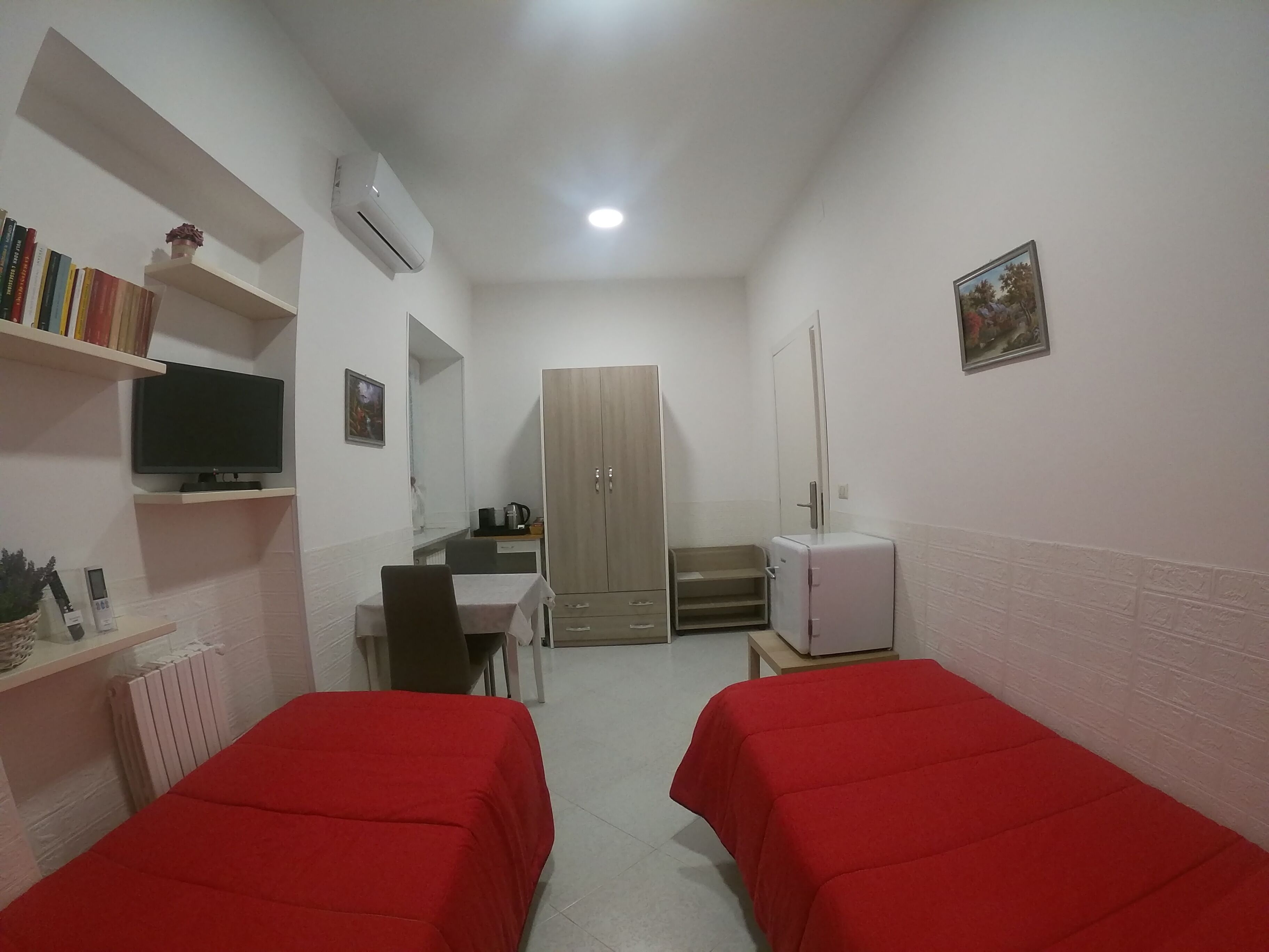 Une petite pièce bien éclairée, comprenant deux lits simples, un bureau avec une chaise, un radiateur, une télévision, une armoire et un réfrigérateur.
