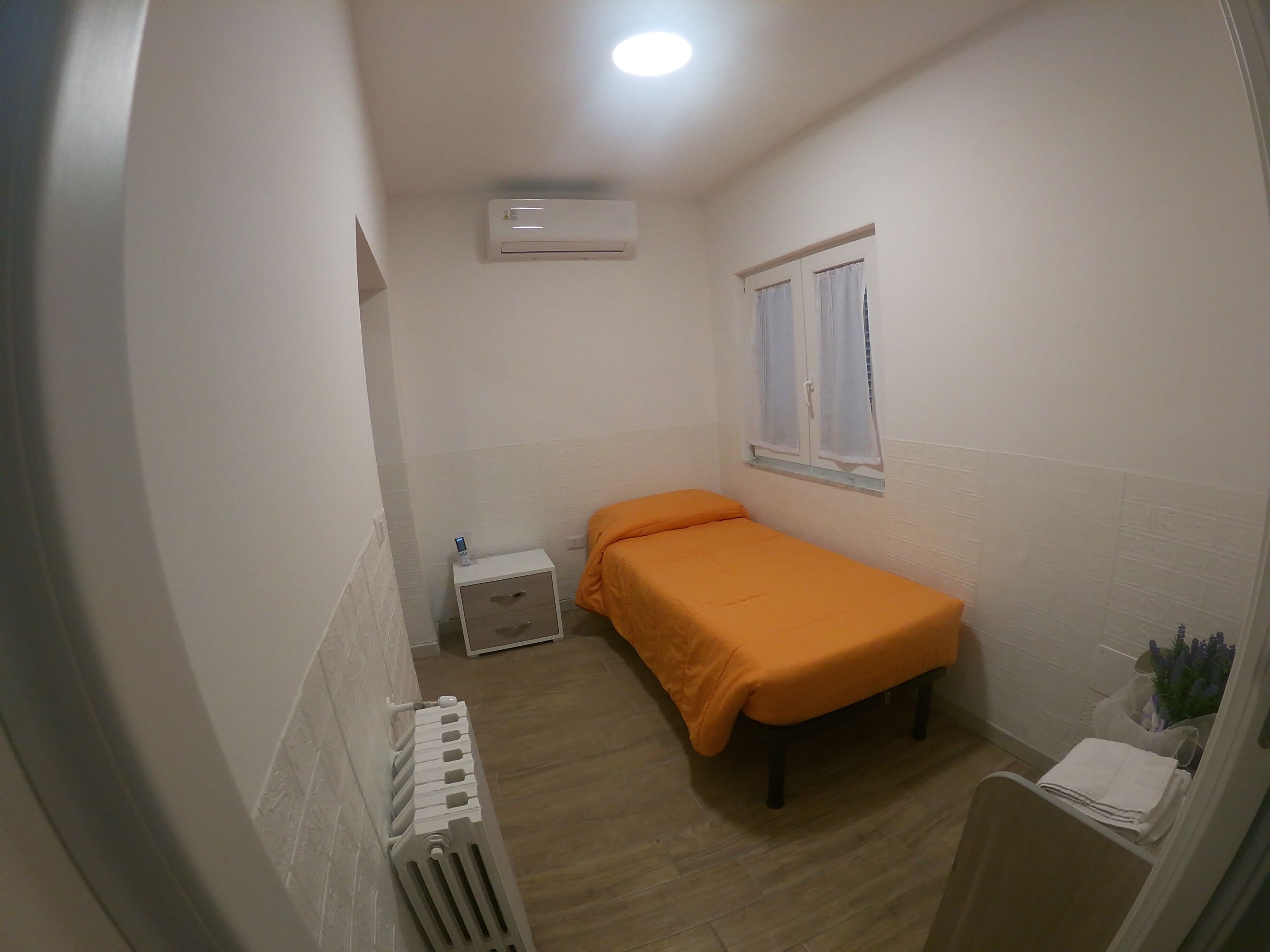 Una habitación pequeña con cama individual, radiador, equipo de aire acondicionado, ventana con cortinas y mesita de noche con lámpara.