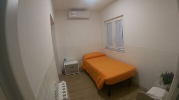 Una habitación pequeña con cama individual, radiador, equipo de aire acondicionado, ventana con cortinas y mesita de noche con lámpara.