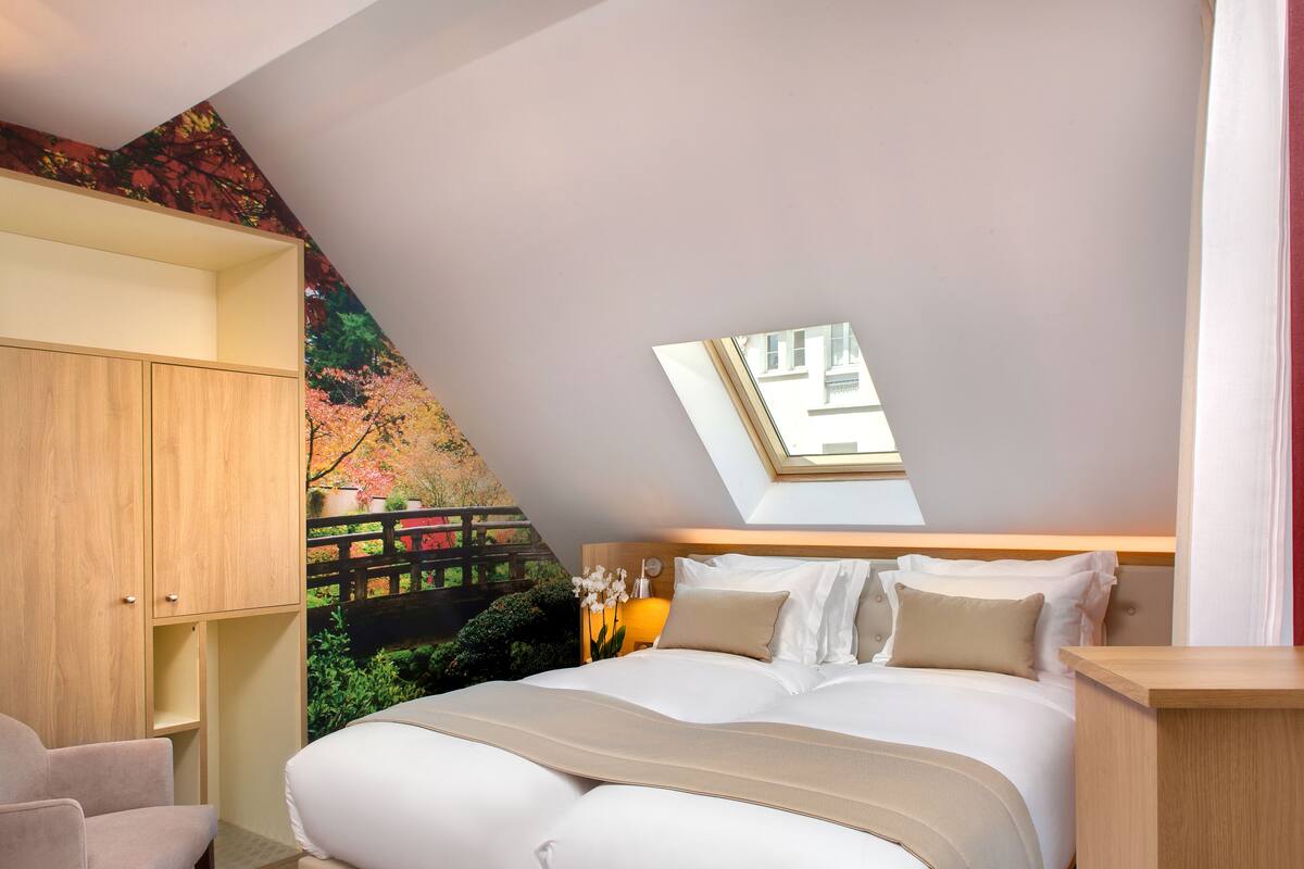 Ein Schlafzimmer mit Dachfenster, einem Bett mit weißer Bettwäsche, einem Holzschrank und einem Wandbild mit Gartenmotiven.