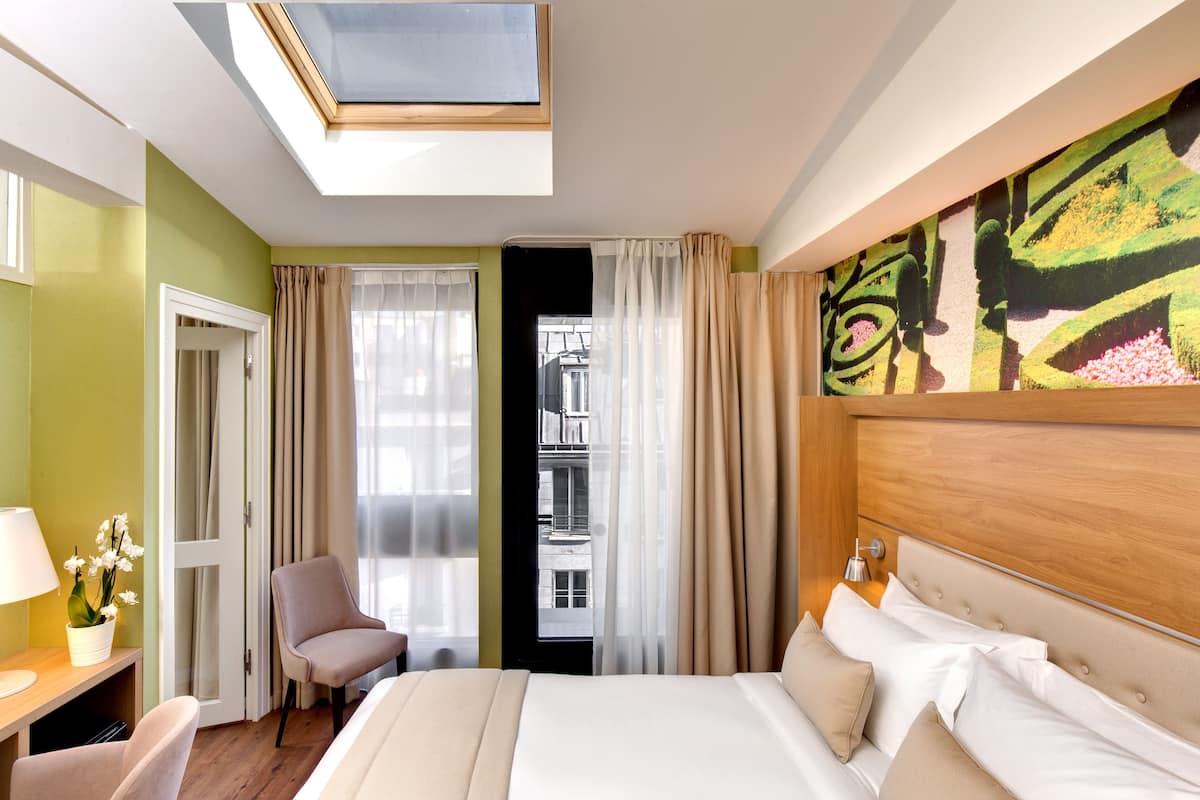 Ein Hotelzimmer mit Bett, Stuhl, Schreibtisch mit Lampe, Fenster mit Vorhängen und einem Oberlicht.
