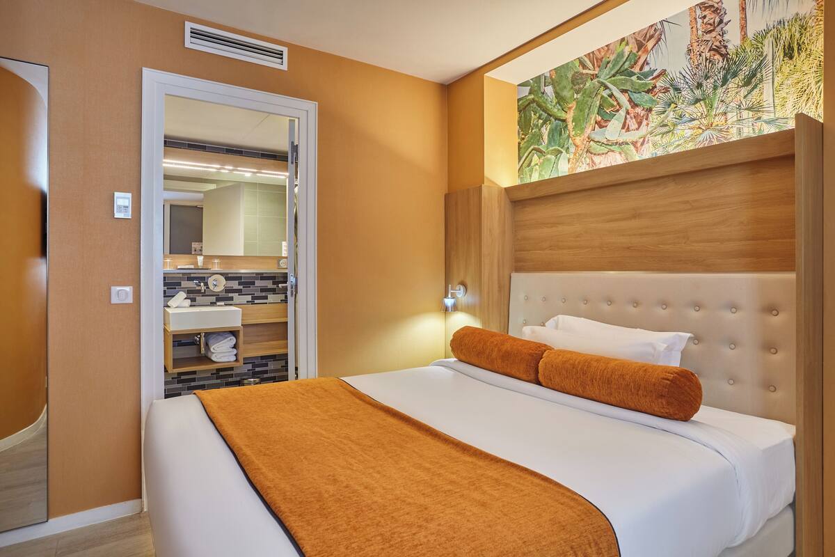 Ein Hotelzimmer mit einem großen Bett, einem Holz-Kopfteil, einem Badezimmer mit Spiegel und Waschbecken sowie einem tropischen Wandbild.