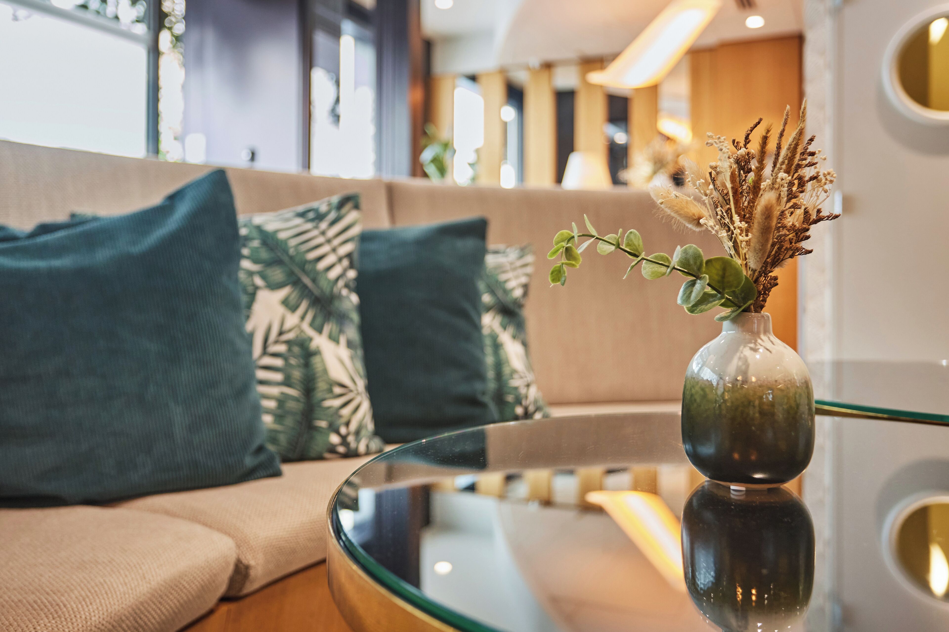 Una sala de estar moderna con una mesa de centro de vidrio, un sofá beige con cojines estampados y un jarr ón decorativo con plantas secas.