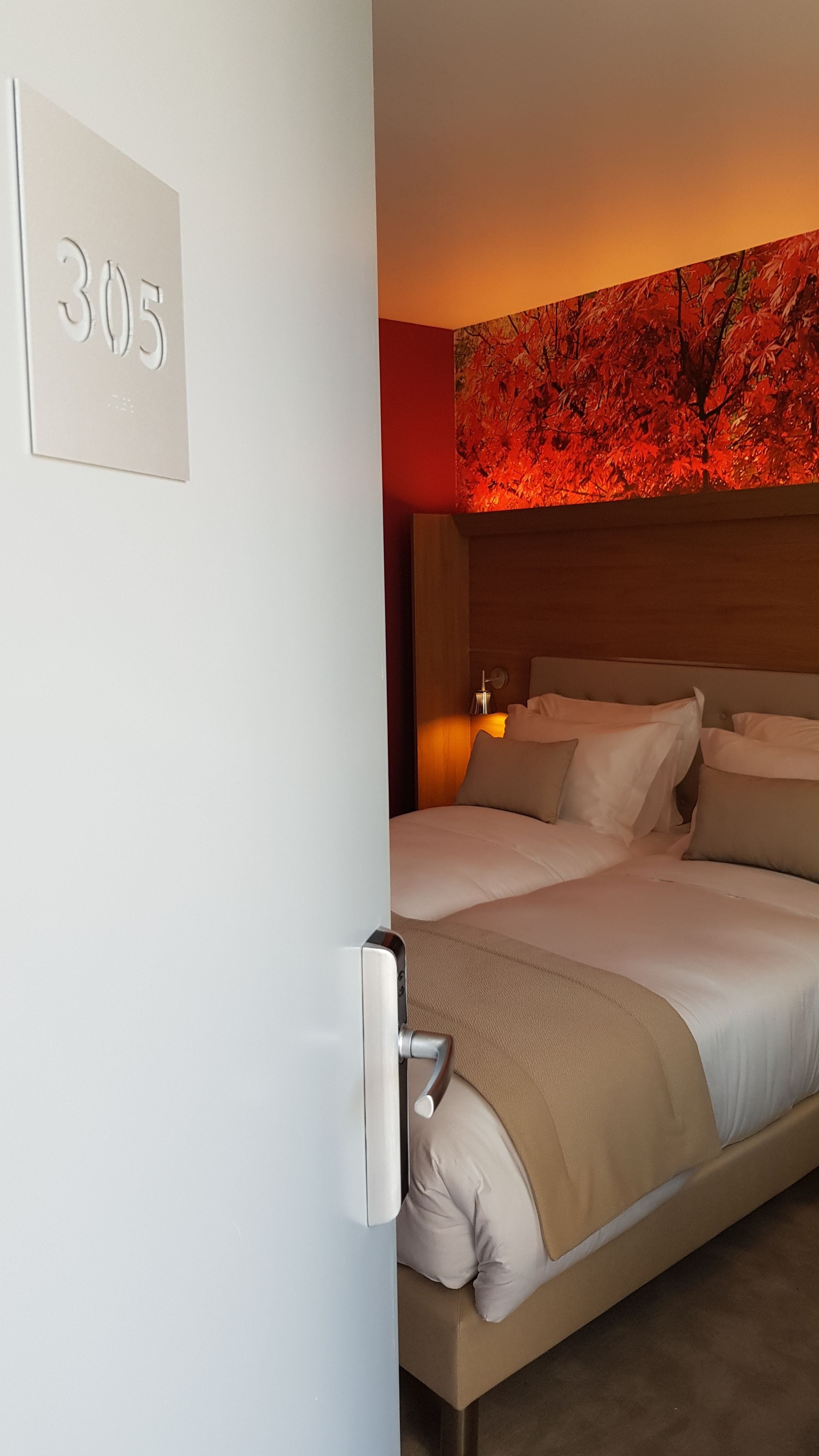 A hotel room with a bed, a nightstand, a wall-mounted lamp, and a number sign.