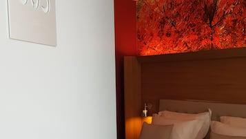 A hotel room with a bed, a nightstand, a wall-mounted lamp, and a number sign.