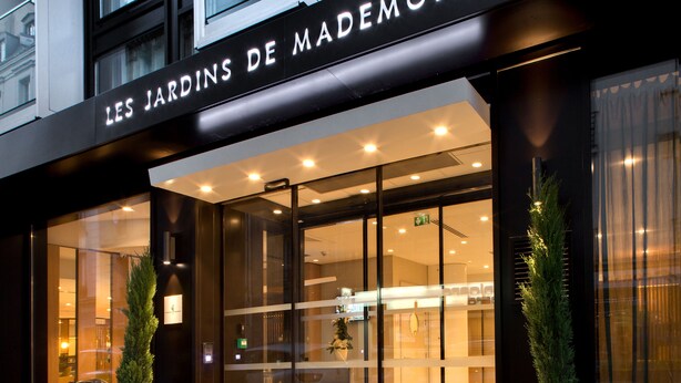 Het uiterlijk van Les Jardins de Mademoiselle, met een moderne gevel, grote glasramen en een opvallend verlicht bord.