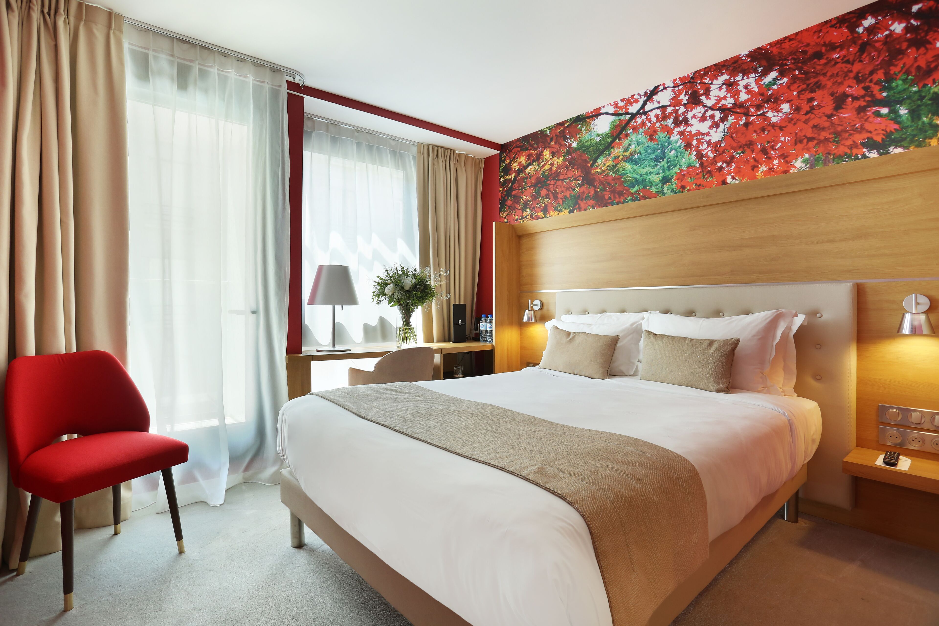 A hotel room with a large bed, a red chair, a desk with a lamp, and a wall with a red and green leaf pattern.