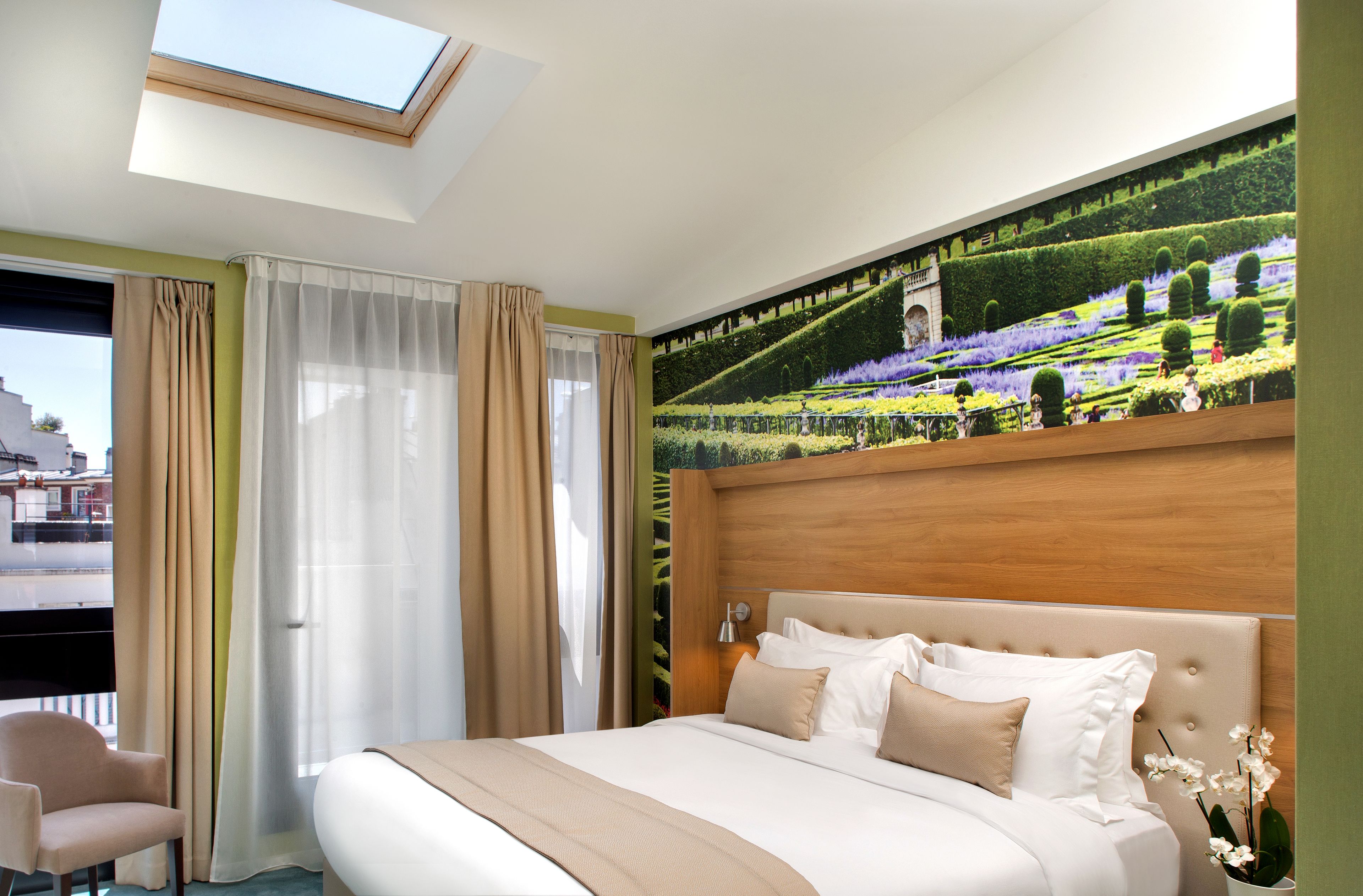 A hotel room with a large bed, a skylight, a mural of a garden, and a view of buildings outside.
