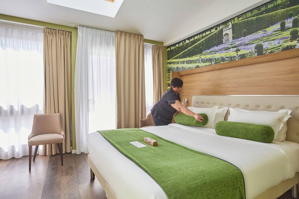 Ein Hotelzimmer mit einem großen Bett, einer grünen Tagesdecke, einem Holzkopfteil und Blick auf einen angelegten Garten durch das Fenster.