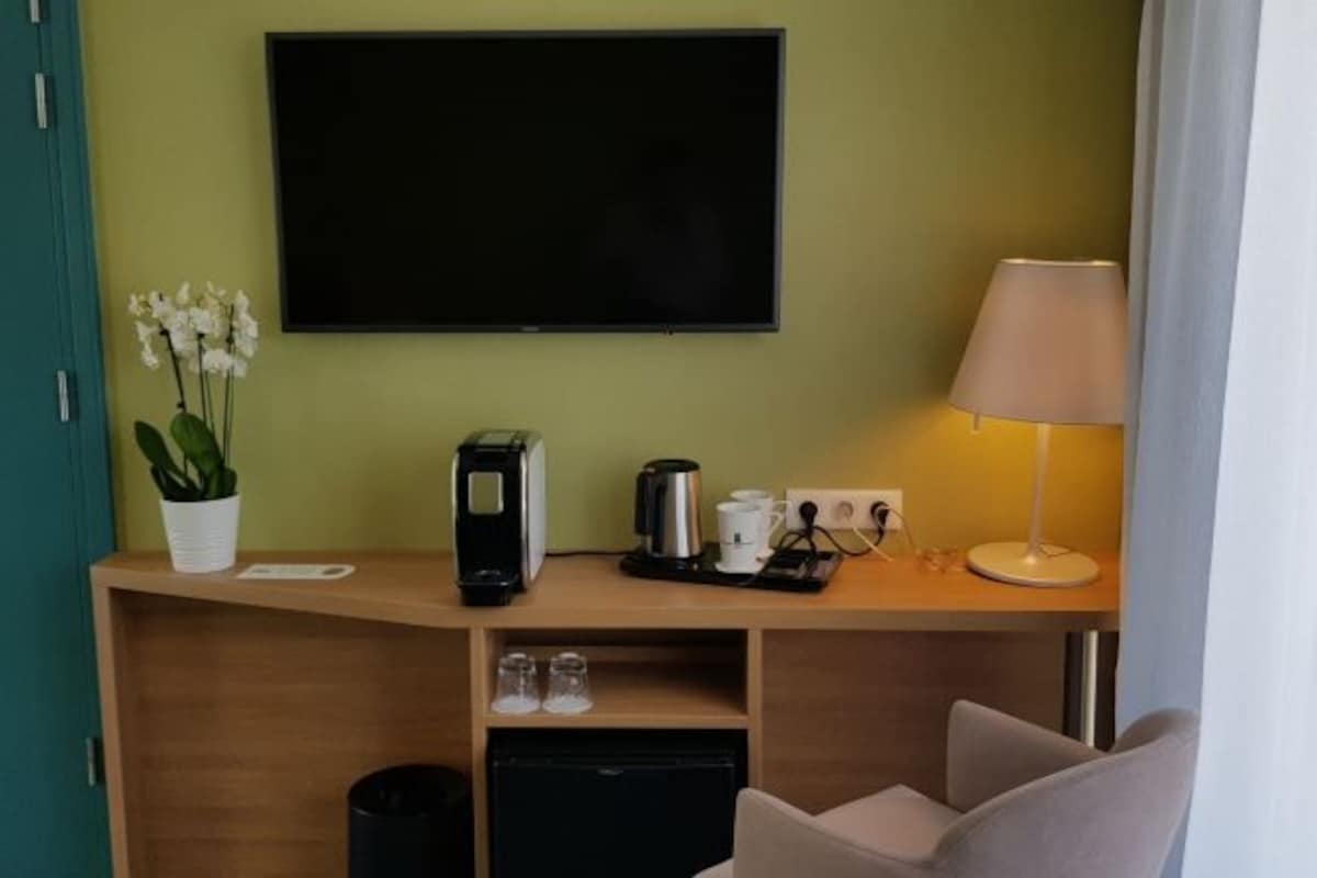 Habitación de hotel con televisor montado en la pared, escritorio con cafetera, lámpara y una silla.