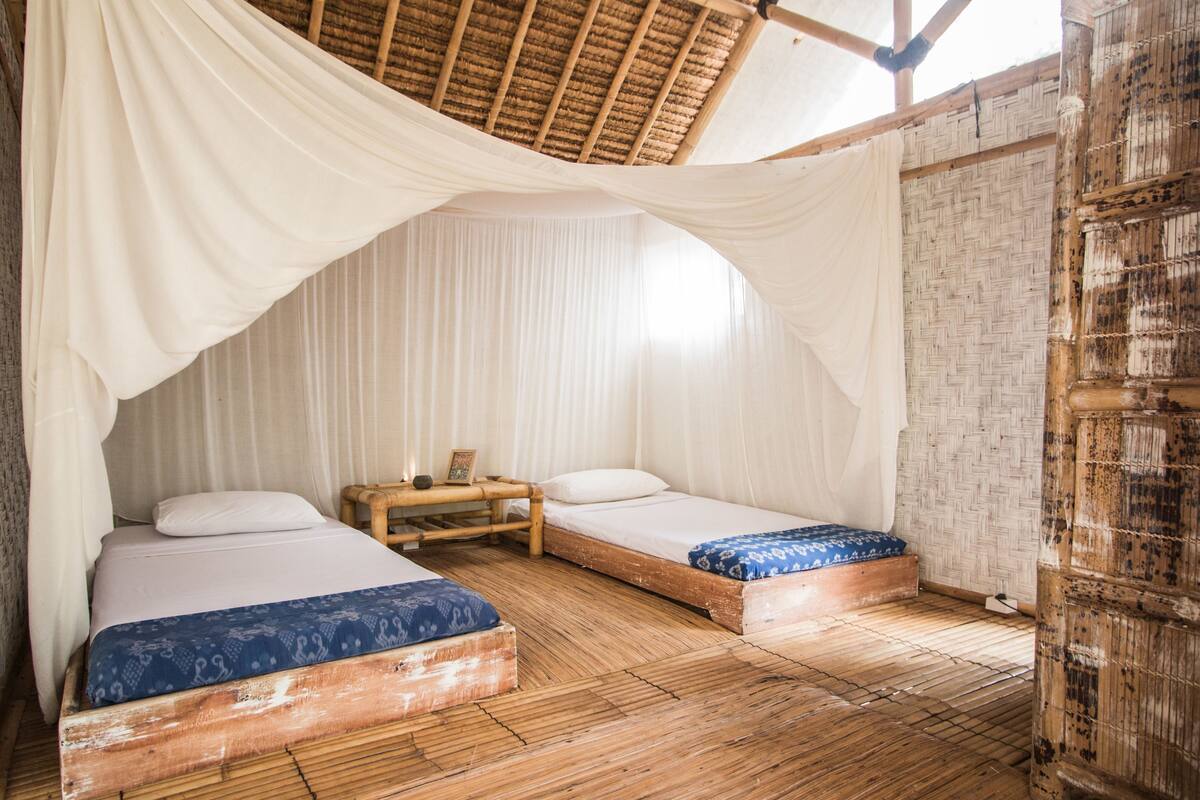 Una habitación con dos camas de madera, cortinas blancas, un banco de madera y un muro tejido.