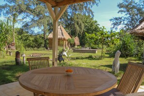 A wooden table with a bowl of fruit in a garden setting with thatched-roof structures and lush greenery. - maoMeno (Gili Meno)