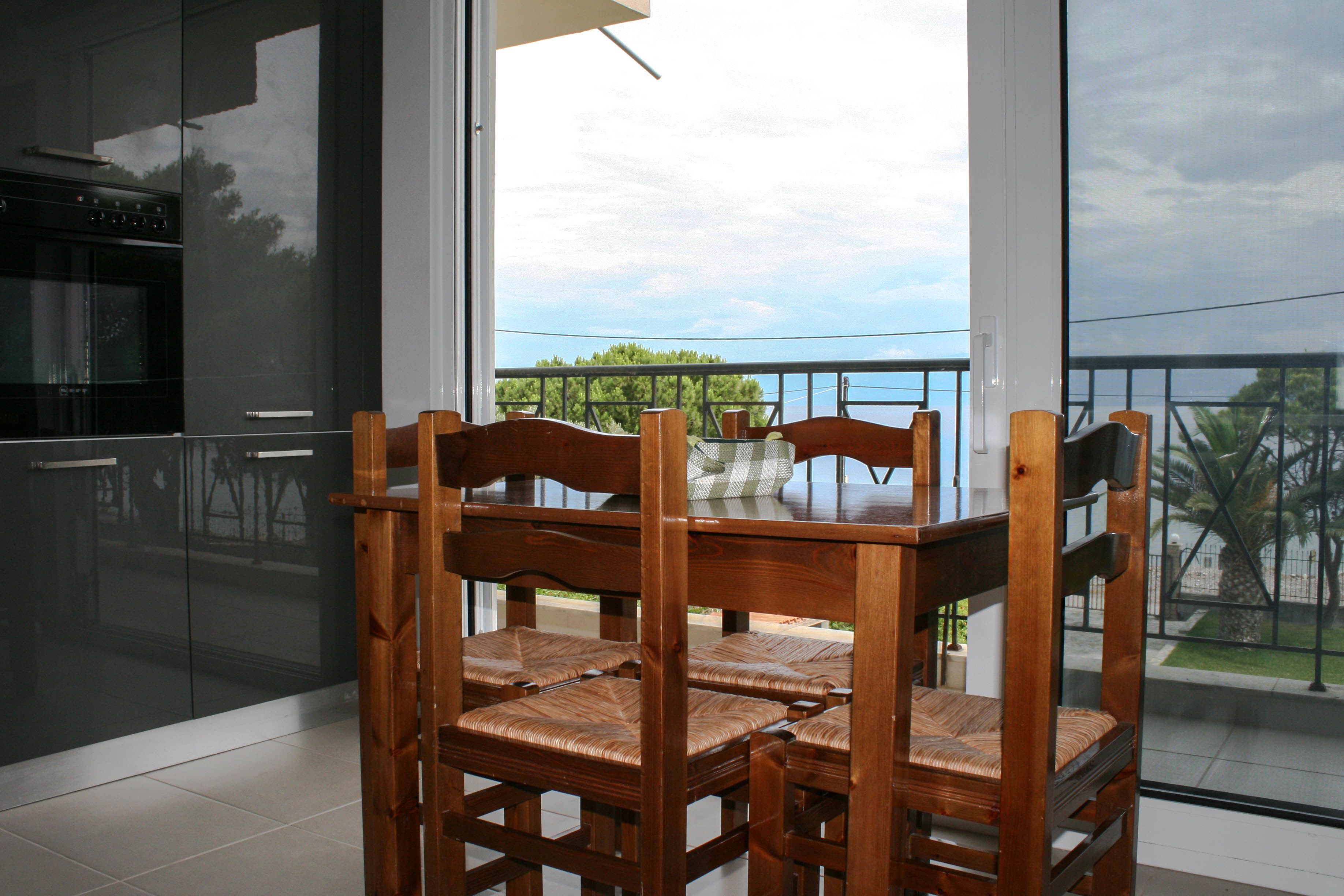 A dining area with a wooden table and chairs, large windows with a view of greenery and a cloudy sky.