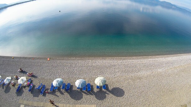 Una playa con agua cristalina, un arenal de guijarros y varios sombrillas y tumbonas.