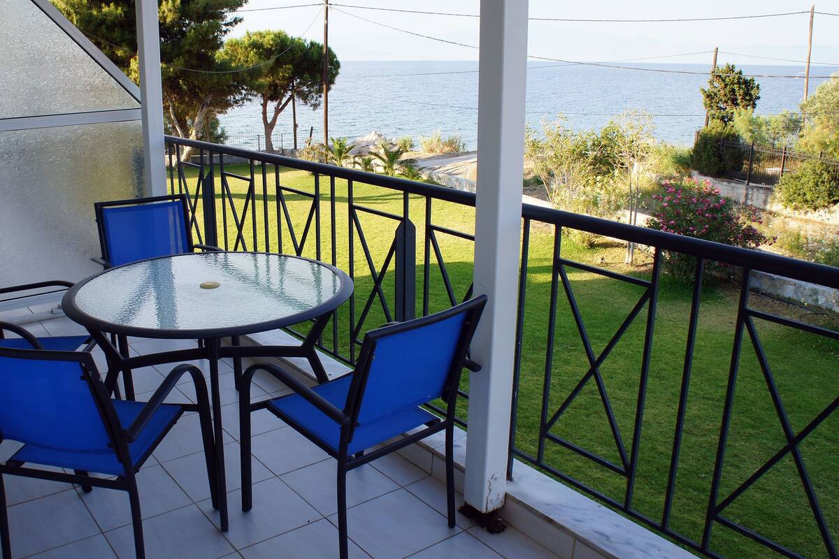 Un balcón con una mesa de vidrio y sillas azules, con vista a un área verde y el mar.