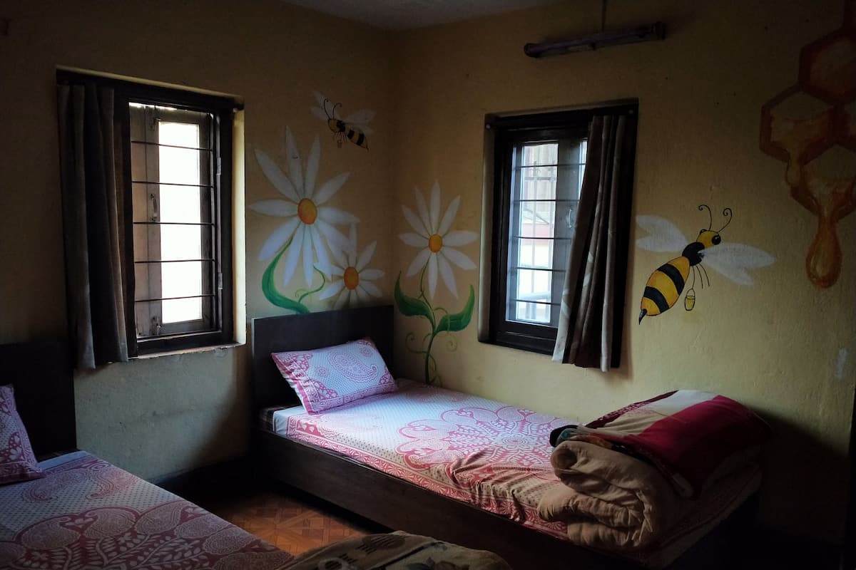A small, dimly lit room with two single beds, a window with curtains, and a wall painted with floral and bee designs.