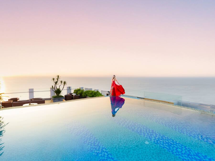 Una persona con un vestido rojo parada junto a una piscina infinita con vistas al océano al atardecer.