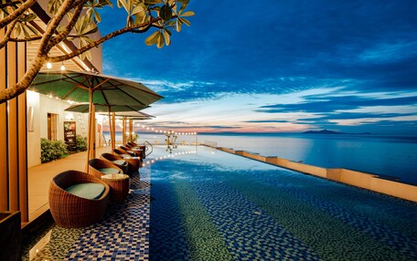 An infinity pool with a checkered floor, wicker chairs with cushions, and a view of the ocean at dusk.