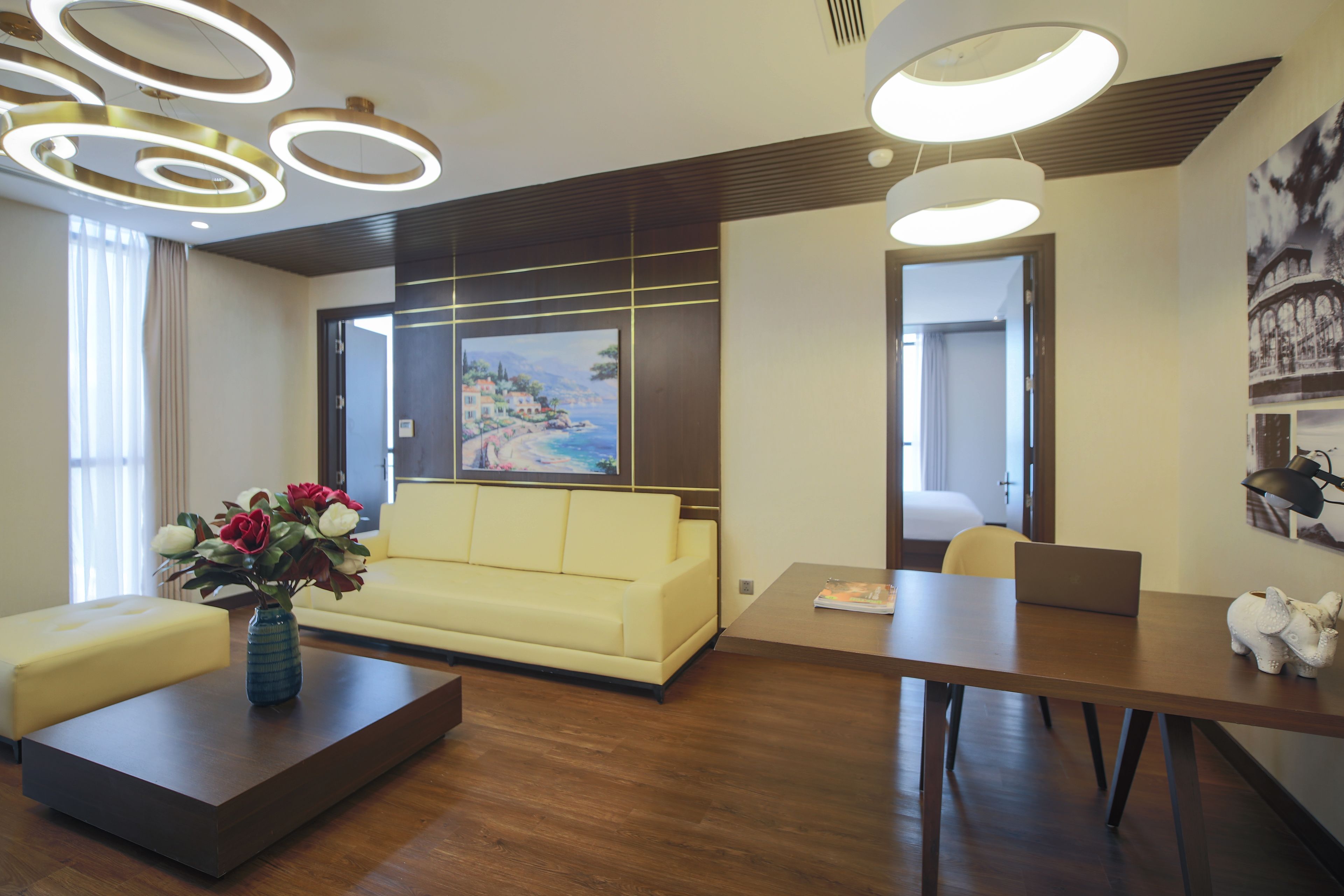 A modern hotel lobby with a yellow sofa, a coffee table with a vase of flowers, a wooden desk with a laptop, and a large painting on the wall.