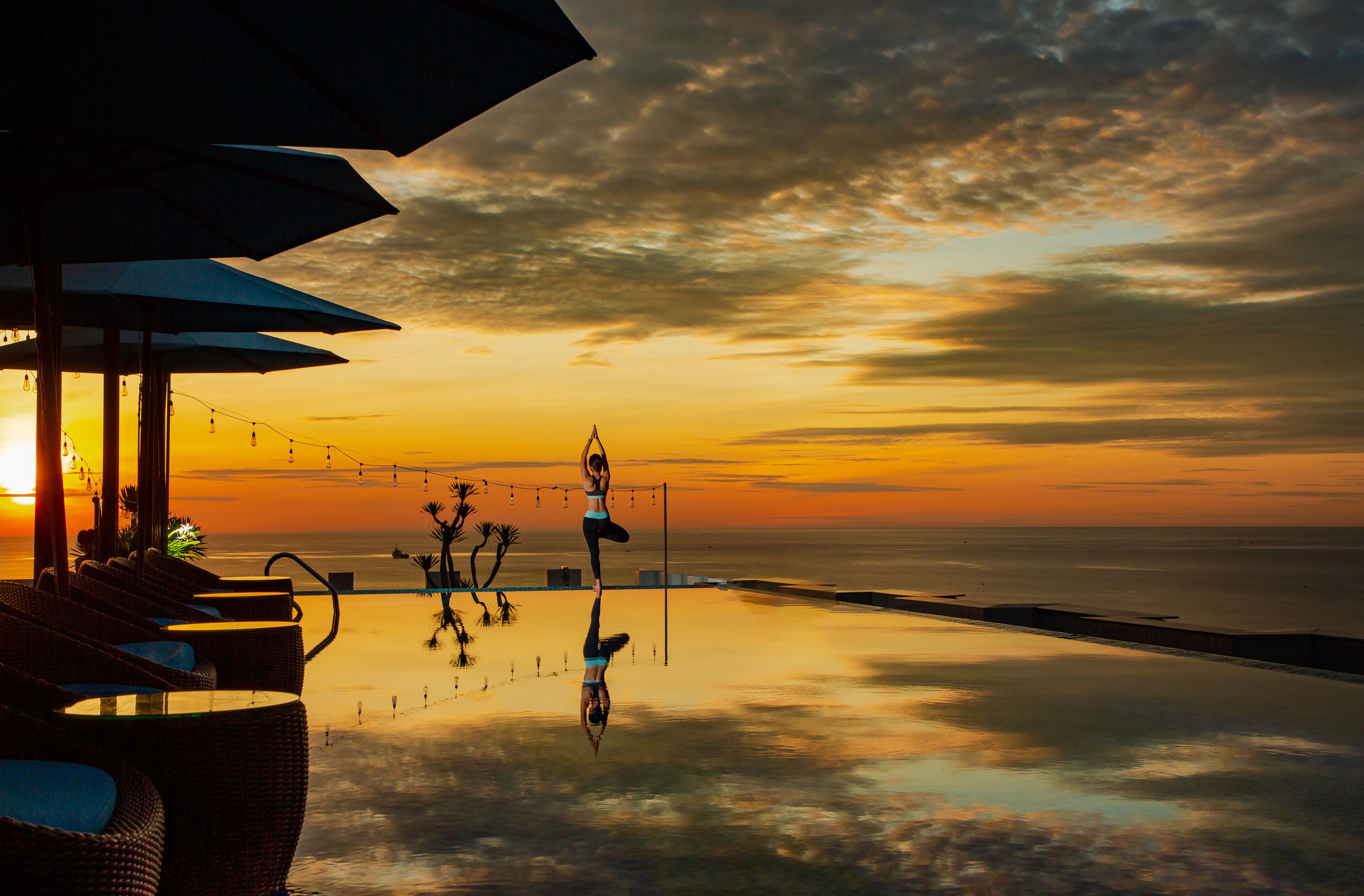 An infinity pool with a view of the sunset over the ocean, outdoor seating with umbrellas, and string lights.