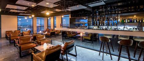 A modern bar area with leather seating, a well-stocked liquor shelf, and large windows offering city views.