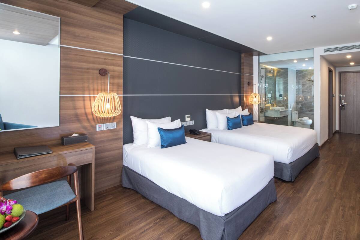 A modern hotel room with two beds, a desk, a chair, a bathroom with a glass shower, and a large mirror.