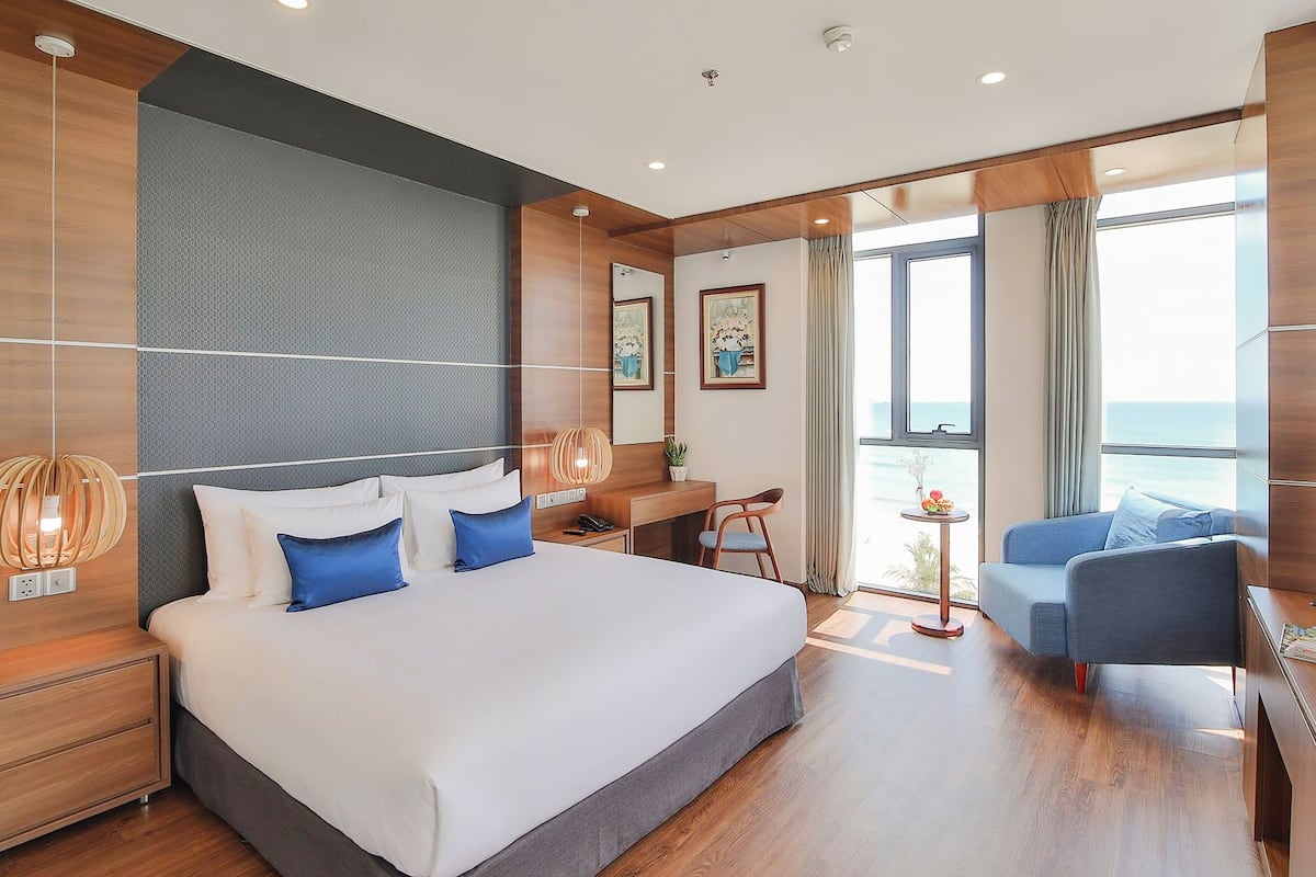 A modern hotel room with a large bed, a desk, a chair, and a view of the sea.