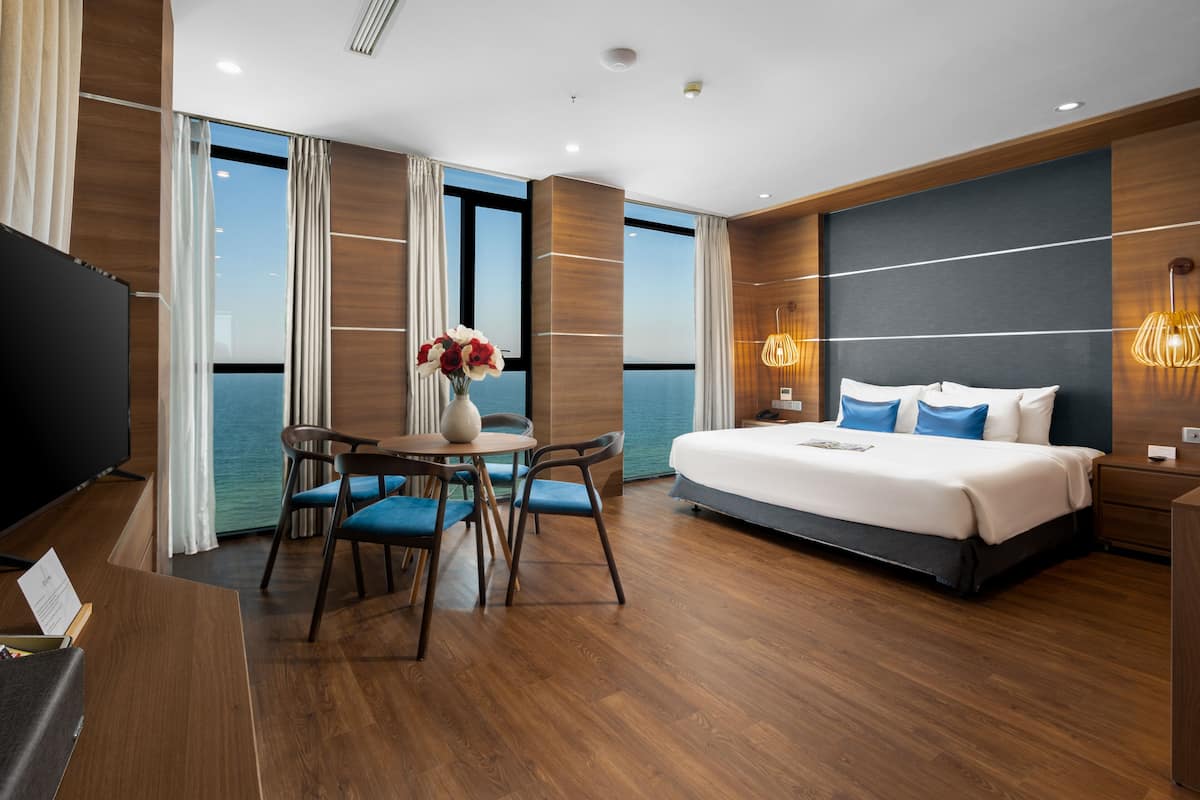 A modern hotel room with a large bed, a desk with a chair, a TV, and a view of the sea through large windows.