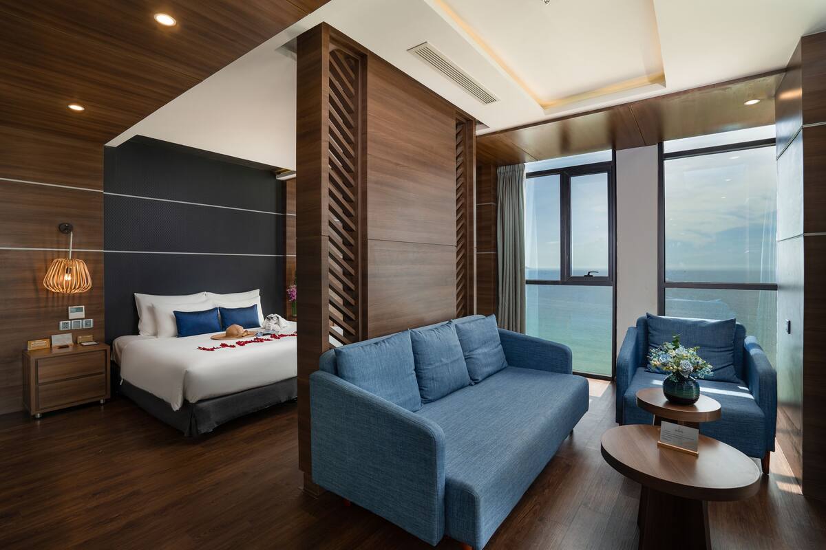 A modern hotel room with a large bed, a blue sofa, and a view of the ocean through large windows.