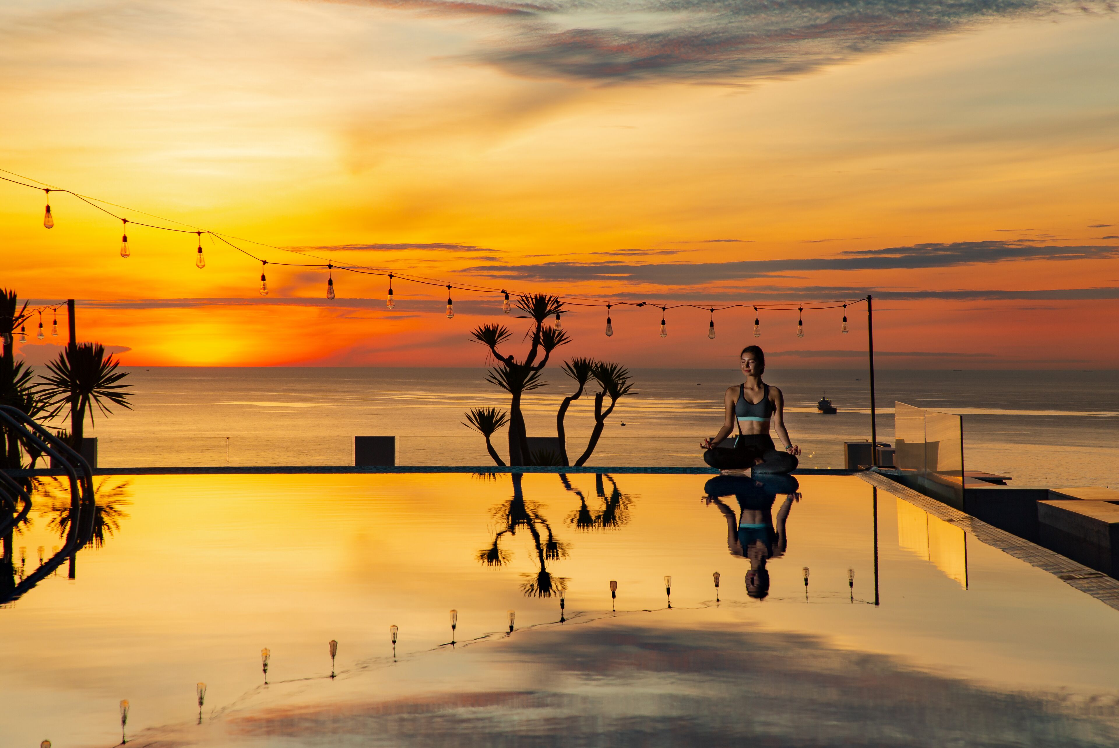 A person meditating on a reflective infinity pool at sunset with string lights and palm trees.