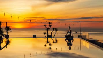 Une personne médite au bord d’une piscine à débordement dont l’eau reflète le ciel au coucher du soleil, le tout illuminé par des guirlandes lumineuses et entouré de palmiers.