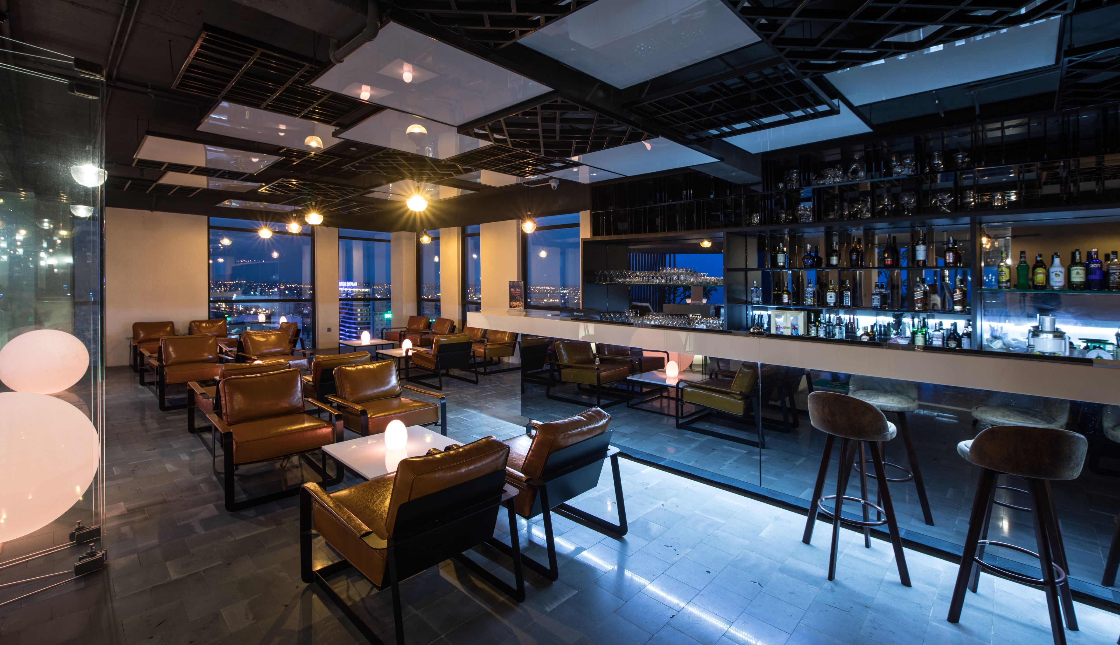 Eine moderne Bar mit Ledersitzgelegenheiten, einer Theke voller Flaschen und großen Fenstern mit Blick auf die Stadt.